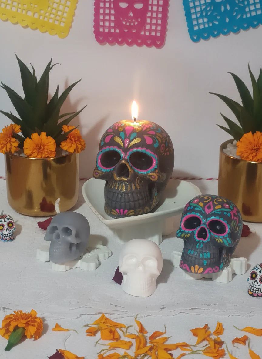 Decorative skull candles and skulls on a table with flowers and colorful paper in the background.